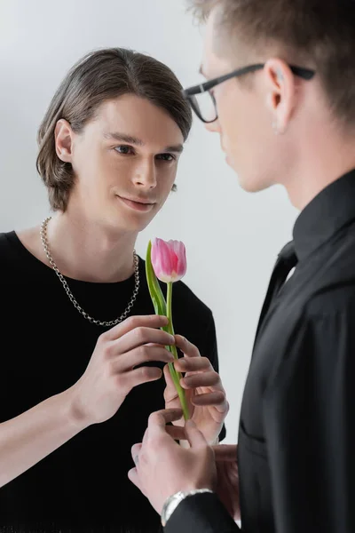 Smiling young gay man holding tulip with blurred boyfriend isolated on grey — Stock Photo