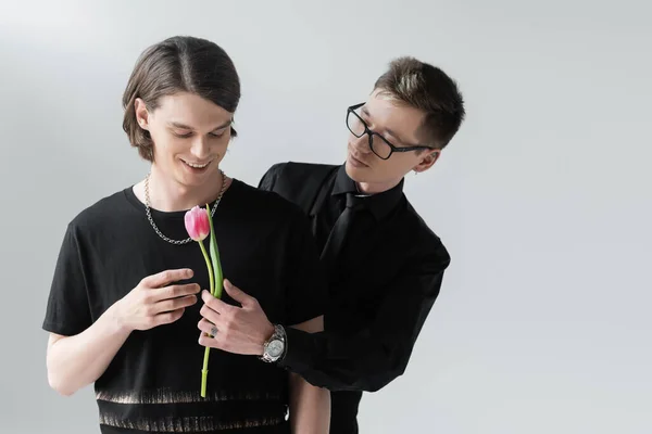 Gay in formal wear and eyeglasses holding flower near positive boyfriend isolated on grey — Stock Photo