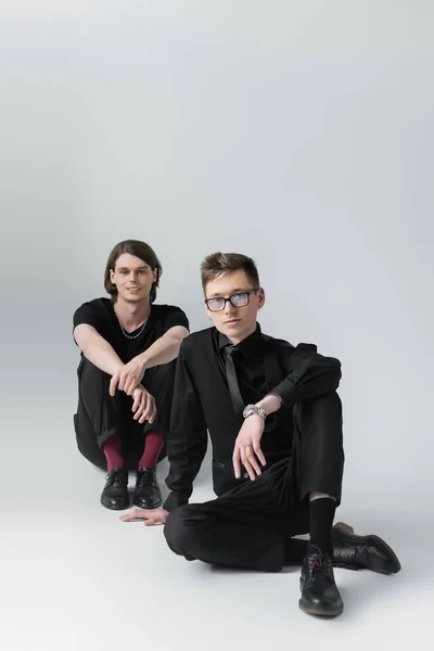 Jeune homme en tenue formelle regardant la caméra tout en étant assis près de petit ami souriant sur fond gris — Photo de stock