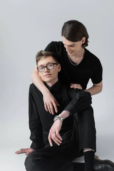 Hombre sonriente abrazando novio en gafas y ropa formal sobre fondo gris - foto de stock