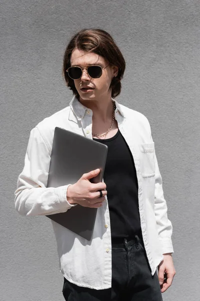 Stylish young man in sunglasses holding laptop outdoors — Stock Photo