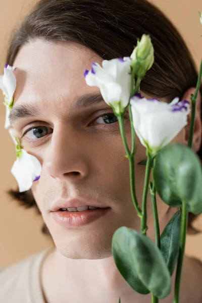 Портрет молодого человека, смотрящего в камеру возле размытого цветка эустомы, изолированного на бежевом — стоковое фото