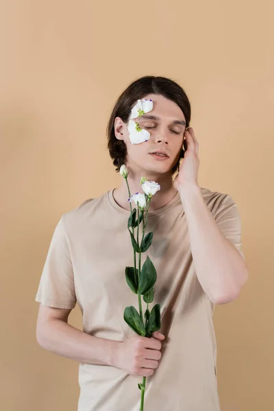 Brunette man holding eustoma and closing eyes isolated on beige — Stock Photo