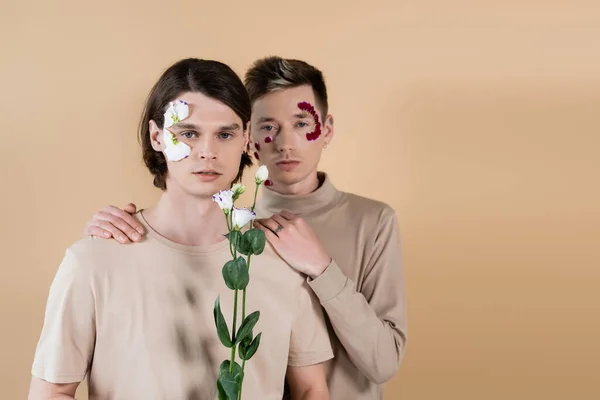 Gay avec pétales sur visage câlin copain avec eustome isolé sur beige — Photo de stock