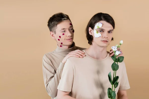 Jeune homme avec des pétales sur le visage câlin partenaire avec eustomie isolé sur beige — Photo de stock