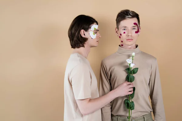 Brunette gay holding eustoma flower near boyfriend with petals on face isolated on beige — Stock Photo