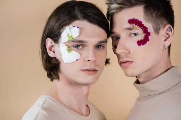 Портрет молодой гомосексуальной пары с лепестками на лицах, смотрящей на камеру, изолированную от бежевого — стоковое фото