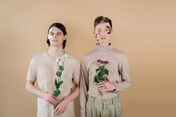 Одностатева пара з пелюстками на обличчях тримає квіти ізольовані на бежевому — стокове фото