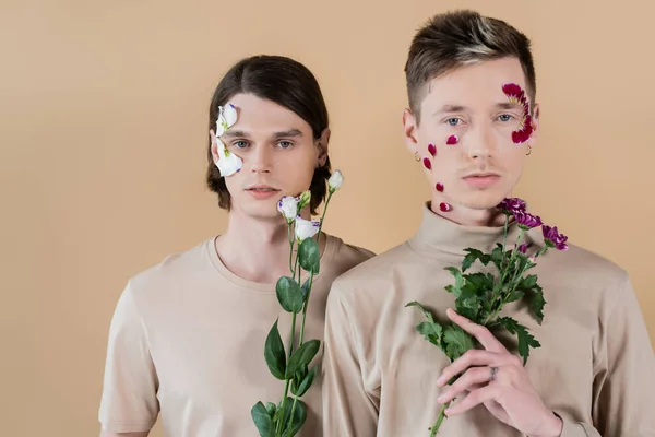 Young gay couple with petals on faces holding flowers isolated on beige — Stock Photo