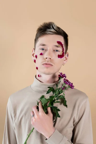 Young man wit petals on face holding chrysanthemums isolated on beige — Stock Photo