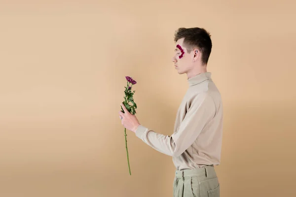 Seitenansicht eines jungen Mannes mit Blütenblättern im Gesicht, der Chrysanthemen isoliert auf Beige betrachtet — Stockfoto