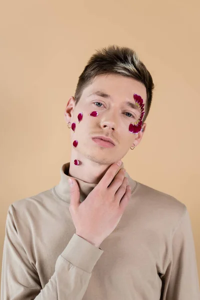 Portrait of young man with petals on face touching neck isolated on beige — Stock Photo