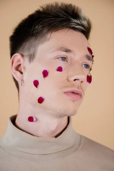 Retrato de un joven con pétalos en la cara mirando hacia otro lado aislado en beige - foto de stock