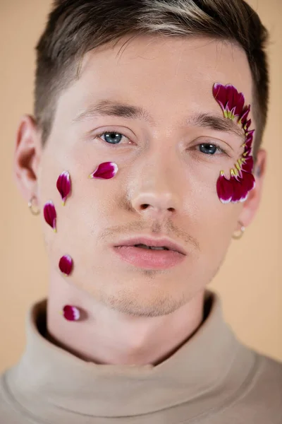 Ritratto di giovane uomo con petali di fiore sul viso guardando la macchina fotografica isolata sul beige — Foto stock