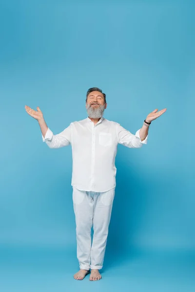 Hombre barbudo descalzo con los ojos cerrados meditando en la pose del árbol con los brazos abiertos en azul - foto de stock