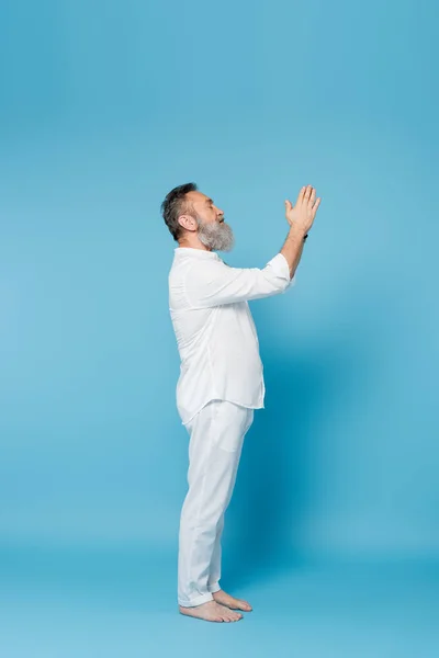 Side view of senior yoga master meditating with praying hands on blue background — Stock Photo