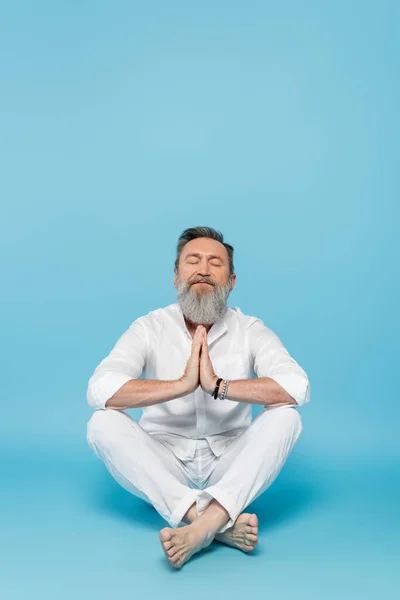 Homem barbudo com olhos fechados e mãos de oração meditando em pose fácil no azul — Fotografia de Stock