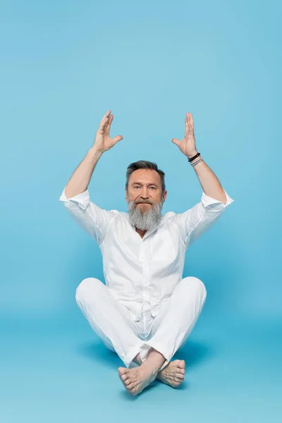 Senior yoga master looking at camera while meditating in easy pose with raised hands on blue — Stock Photo