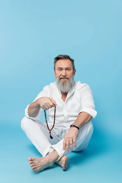 Bearded barefoot man sitting with crossed legs and holding mala beads on blue background — Stock Photo