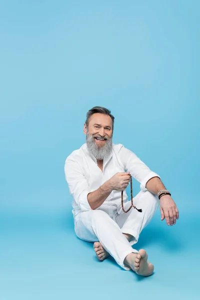 Cheerful yoga master sitting with crossed legs and mala beads on blue background — Stock Photo