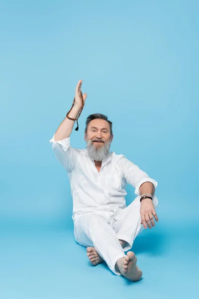 Smiling guru teacher sitting with crossed legs and mala beads in raised hand on blue — Stock Photo