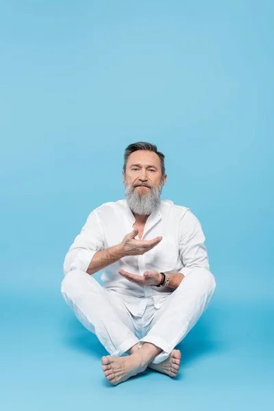 Bearded man in white clothes meditating in easy pose on blue background — Stock Photo