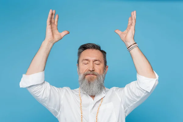 Gurú barbudo hombre meditando con los ojos cerrados y las manos levantadas aisladas en azul - foto de stock