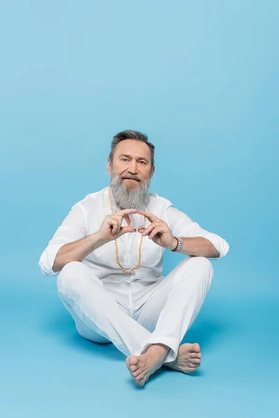 Guru tutor sênior mostrando hakini mudra enquanto sentado com pernas cruzadas em azul — Fotografia de Stock