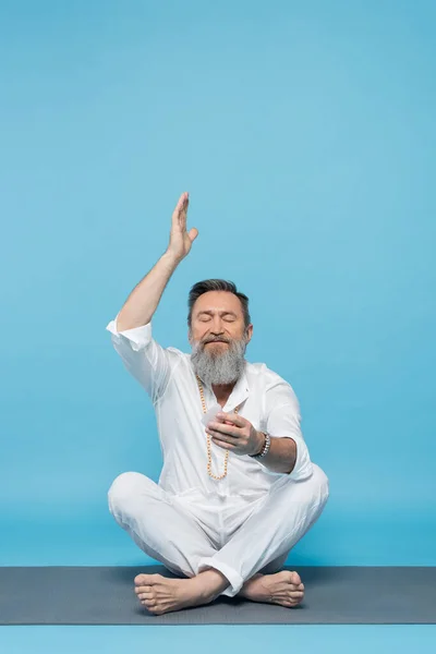 Uomo anziano in abiti bianchi meditando con cristallo selenite in posa facile con mano sollevata sul blu — Foto stock