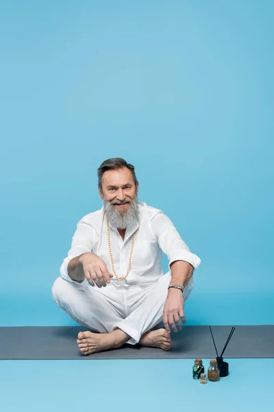 Maestro de yoga feliz sentado en pose fácil cerca de aceites esenciales y palitos de aroma en azul - foto de stock