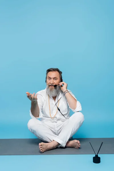 Yoga coach in headset sitting in easy pose on yoga mat and pointing with hand on blue — Stock Photo