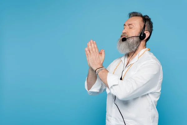 Hombre gurú sénior en auriculares meditando con los ojos cerrados y las manos rezando aisladas en azul - foto de stock