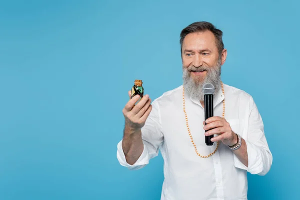 Smiling ayurveda guru with bottle of essential oil talking in microphone isolated on blue — Stock Photo