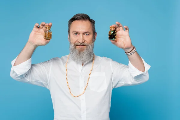 Guru de cura barbudo segurando frascos com óleos perfumados e olhando para a câmera isolada em azul — Fotografia de Stock