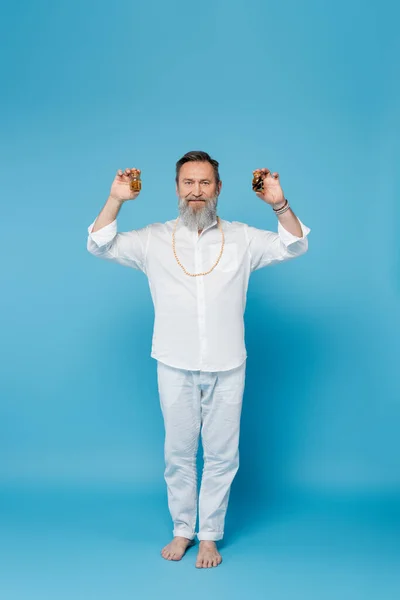 Barefoot bearded man in white clothes holding bottles with essential oils on blue — Stock Photo