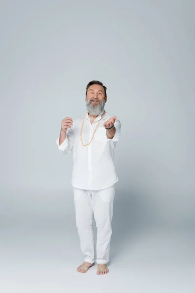 Pleine longueur de coach de yoga heureux en vêtements blancs regardant la caméra et pointant avec la main sur le gris — Photo de stock