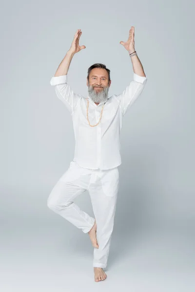 Full length of senior yoga coach meditating in tree pose with raised hands on grey — Stock Photo