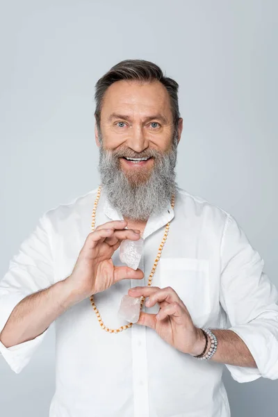 Senior spiritual coach holding crystals of selenite while smiling at camera isolated on grey — Stock Photo