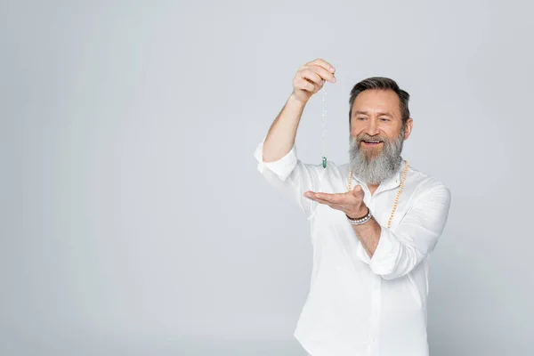 Anziano guru uomo sorridente mentre medita con pendolo rabdomante isolato su grigio — Foto stock