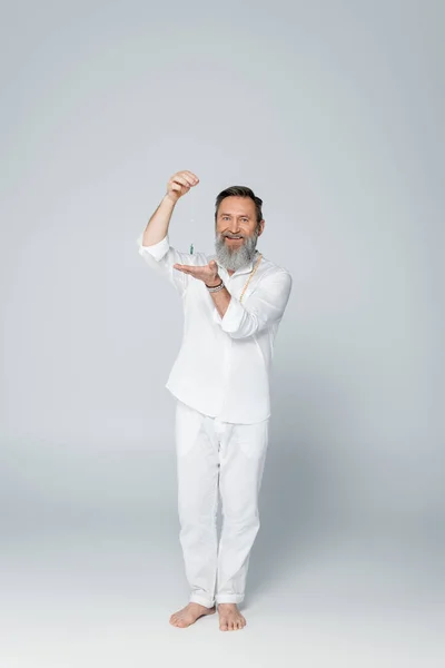 Full length of happy yoga coach with dowsing pendulum looking at camera on grey — Stock Photo