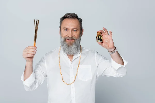 Ayurveda guru with aroma sticks and scented oil smiling at camera isolated on grey — Stock Photo