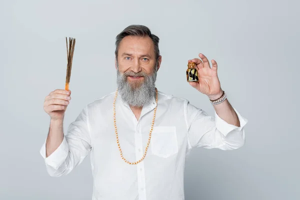 Ayurveda-Guru mit Aromastäbchen und duftendem Öl lächelt isoliert auf grau in die Kamera — Stockfoto