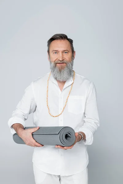 Guru mestre barbudo em camisa branca e contas segurando tapete de ioga isolado em cinza — Fotografia de Stock