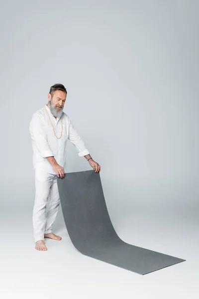 Full length of senior barefoot man in white clothes holding yoga mat on grey — Stock Photo