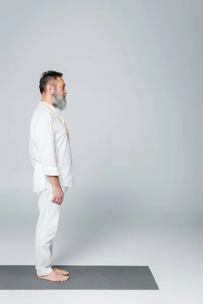 Vue latérale de l'entraîneur de yoga barbu méditant en pose de montagne sur tapis de yoga sur gris — Stock Photo