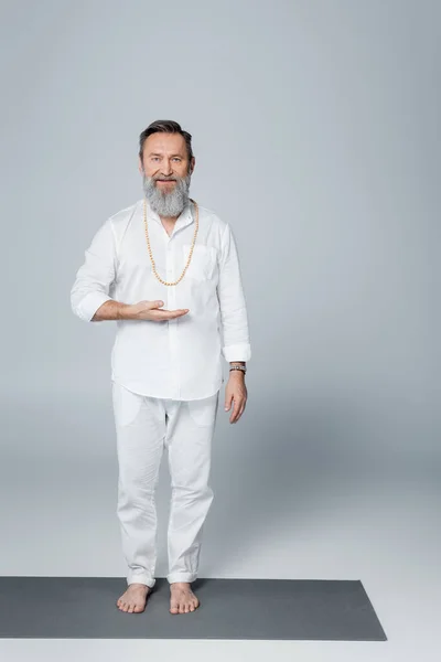 Senior barefoot spiritual guru pointing with hand and looking at camera on grey — Stock Photo