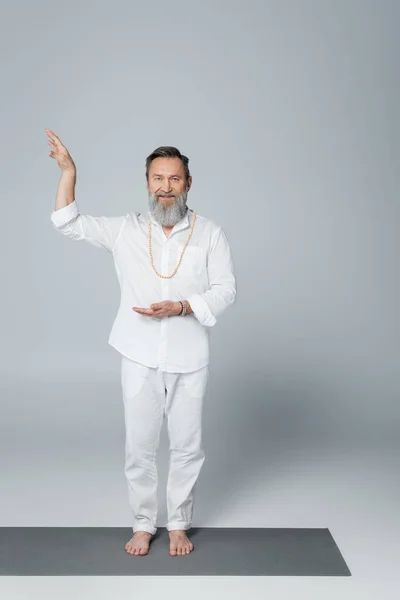 Bearded and barefoot guru man in white clothes gesturing and looking at camera on grey — Stock Photo