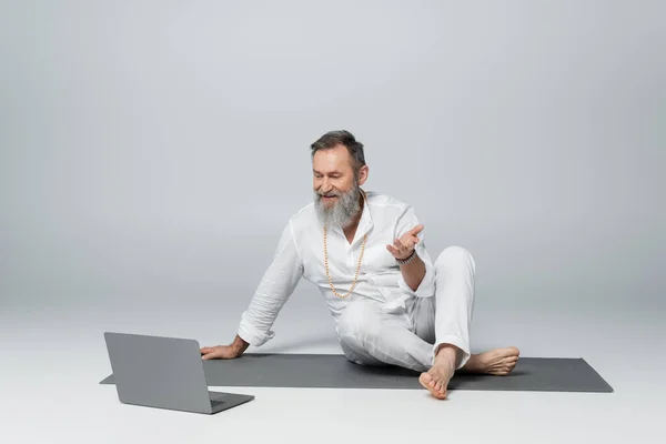 Mentor gurú sentado en esterilla de yoga con las piernas cruzadas y hablando cerca del ordenador portátil en gris - foto de stock