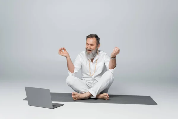 Grey haired yoga master sitting in easy pose and showing chin mudra near laptop on grey — Stock Photo