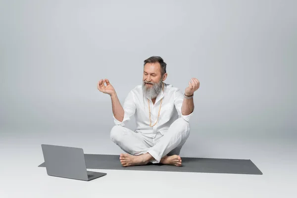 Maestro di yoga dai capelli grigi seduto in posa facile e mostrando mudra mento vicino laptop su grigio — Foto stock